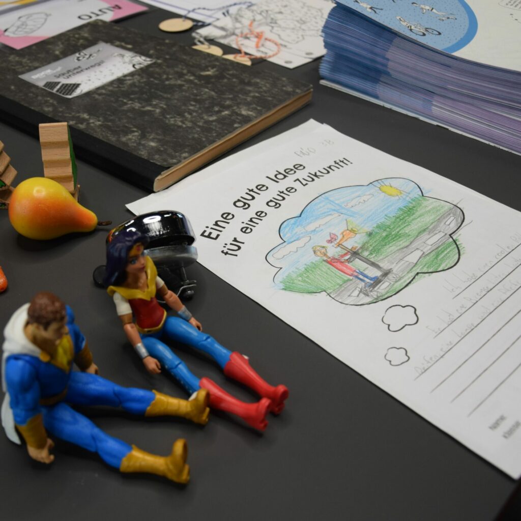 2 Spielfiguren sitzen auf einem Tisch, daneben liegt ein Plakat mit dem Titel: Eine gute Idee für eine gute Zukunft, darunter sind Zeilen, die zum Ausfüllen mit Stiften einladen.