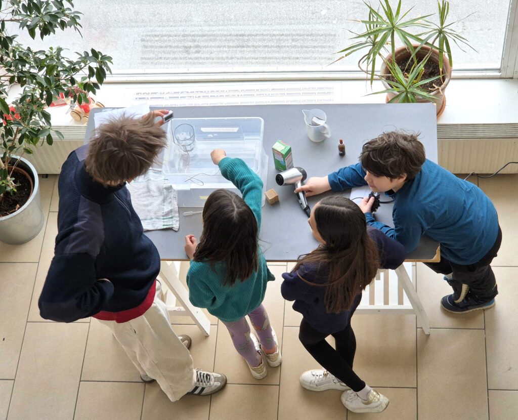 3 Kinder und ein Erwachsenen vor einem Wasserbehälter mit Messstreifen, experimentieren